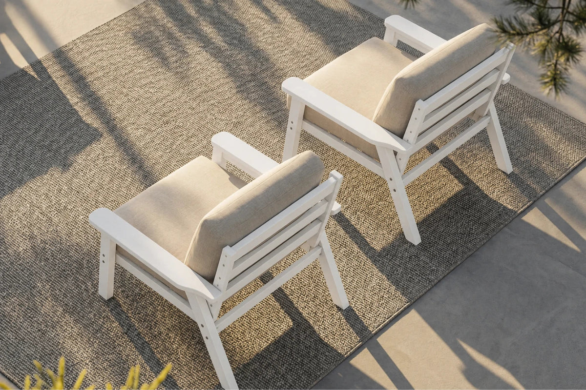 Two white outdoor armchairs with creamy white cushions arranged on a textured gray rug over a concrete patio, with dappled sunlight and shadows.