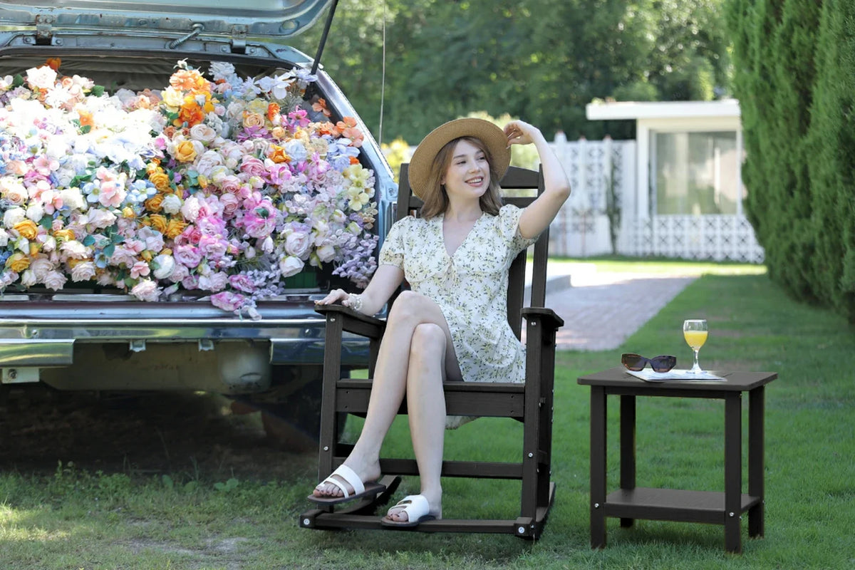 Woman sitting in a rocking chair next to a truck filled with flowers, with a table and sunglasses in the foreground.