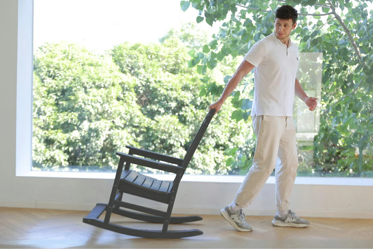 Person pushing a black rocking chair indoors with large windows showing greenery outside.
