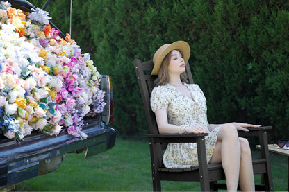 Woman in a floral dress sitting on a wooden chair next to a truck with flowers, against a green hedge background.