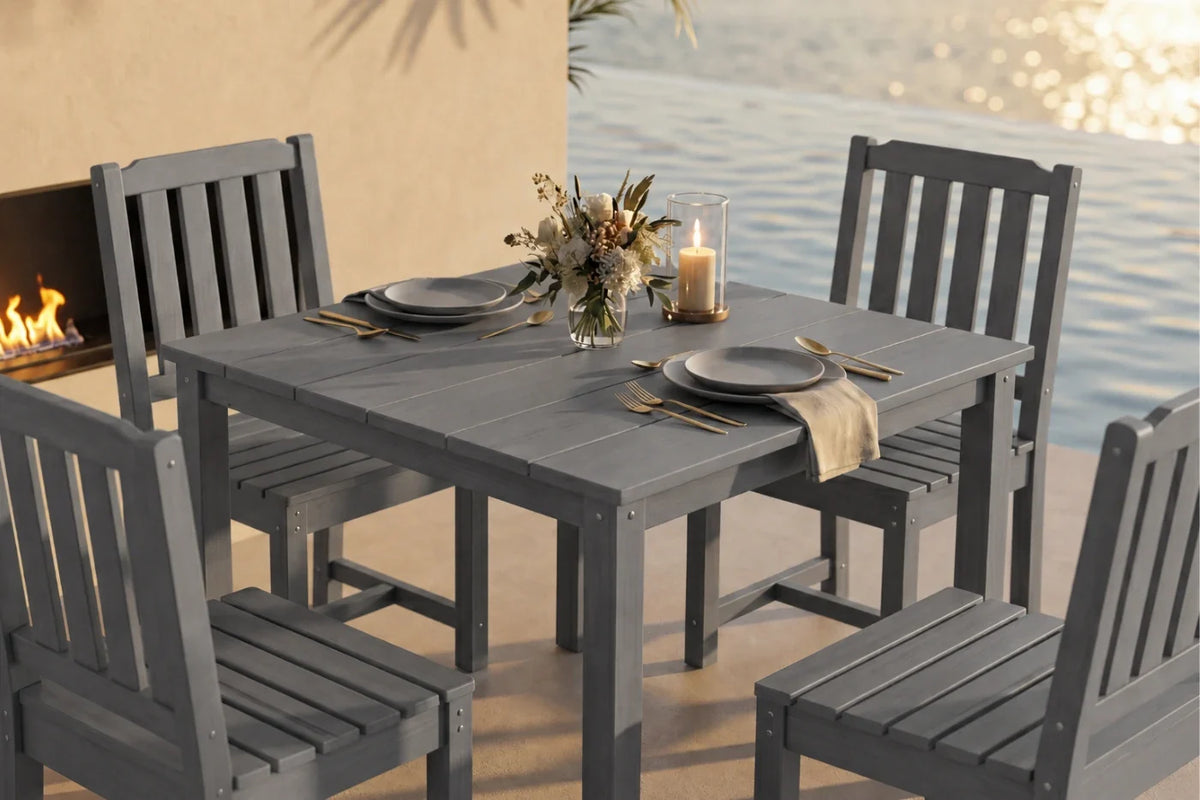 A 5-piece gray outdoor dining set consisting of a  table and four matching armless chairs, styled with plates, cutlery, a candle, and a floral centerpiece on a patio with a fireplace and water in the background.
