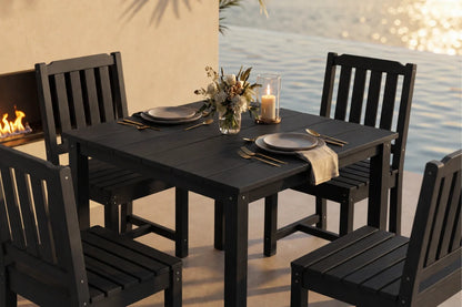 A black outdoor dining set consisting of a  table and four matching armless chairs, styled with plates, cutlery, a candle, and a floral centerpiece on a patio with a fireplace and water in the background.