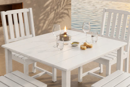 White outdoor dining table with 4 armless chairs, glasses, and a candle on a patio.