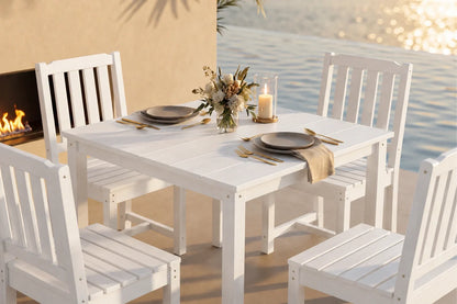 A white outdoor dining set consisting of a  table and four matching armless chairs, styled with plates, cutlery, a candle, and a floral centerpiece on a patio with a fireplace and water in the background.