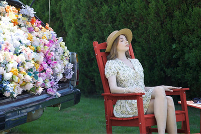 Woman in a floral dress sitting on a red chair next to a truck with flowers, against a green hedge background.