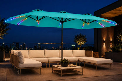 A large outdoor patio umbrella with glowing aruba blue fabric and multicolor RGB LED light strips along the edges, shading a beige sectional sofa and coffee table on a rooftop terrace at night.