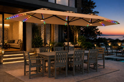 A large beige patio umbrella with colorful RGB LED string lights along the canopy, shading a wooden dining table and chairs on a patio at dusk. A modern house, pool area, and sunset are visible in the background.