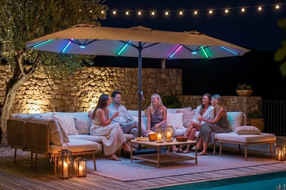 Outdoor patio scene at night with a large beige patio umbrella illuminated by colorful RGB LED strip lights along the canopy edge. Under the umbrella, a group of five people relax on a light beige sectional sofa and chaise lounge pieces around a low wooden coffee table with candles.