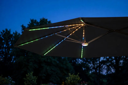 A dark patio umbrella outdoors at dusk with RGB LED string lights running along the canopy and radiating from the center hub, casting green and blue light beams against a deep blue evening sky and silhouetted trees.