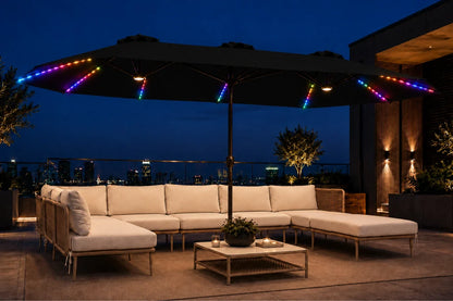 Outdoor patio furniture set under a large black patio umbrella at dusk, featuring a sectional sofa, matching chaise, low coffee table, and multicolor RGB LED string lights along the umbrella canopy and railings.