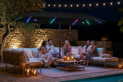 Outdoor patio scene at night with a group of five people sitting on a beige sectional sofa under a large black patio umbrella. The umbrella is illuminated with multicolor RGB LED light strips along the canopy edge, casting colored glows onto the surrounding stone wall and deck.