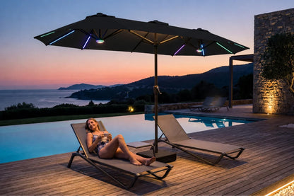 A woman relaxes on a beige lounge chair beside an outdoor pool at dusk, with a large black patio umbrella featuring integrated multicolor RGB LED light strips overhead. A second lounge chair sits nearby, and a stone wall and distant hills are visible in the background.