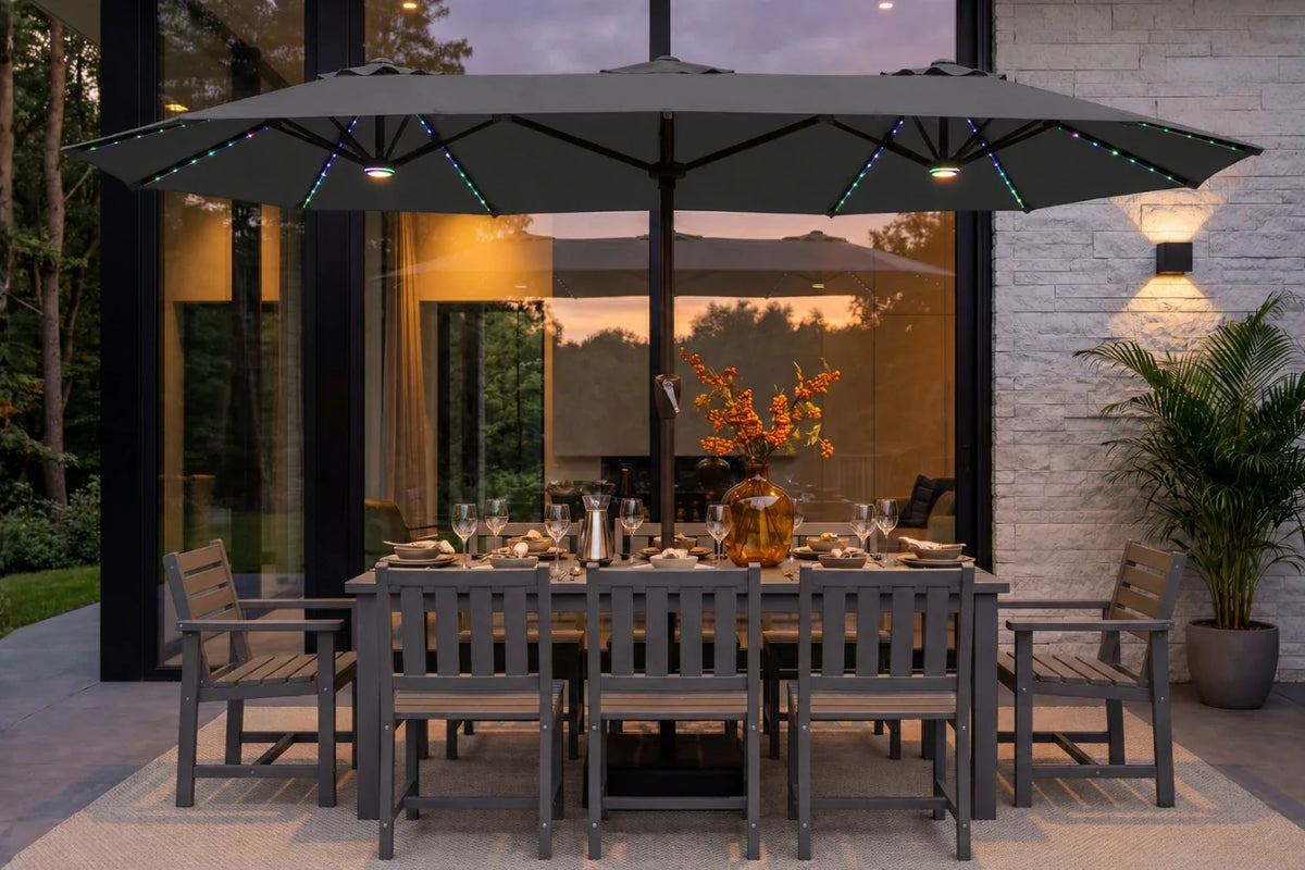 A large dark gray outdoor patio umbrella with integrated RGB LED string lights is shown over a matching gray dining table and chairs on a patio in front of a modern house at dusk.