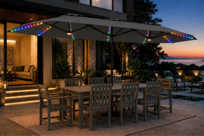 A large dark gray outdoor patio umbrella with a matching dining table and chairs on a patio at dusk, decorated with multicolor RGB LED string lights along the canopy and frame.