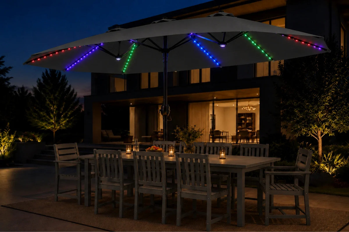 A large dark gray patio umbrella with colorful RGB LED string lights along its edge, shading an outdoor dining table and chairs in front of a modern house at night.