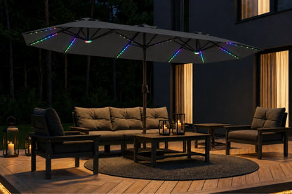 Outdoor patio furniture set under a large dark gray patio umbrella with multicolor RGB LED string lights, shown at night on a wooden deck beside a modern house.