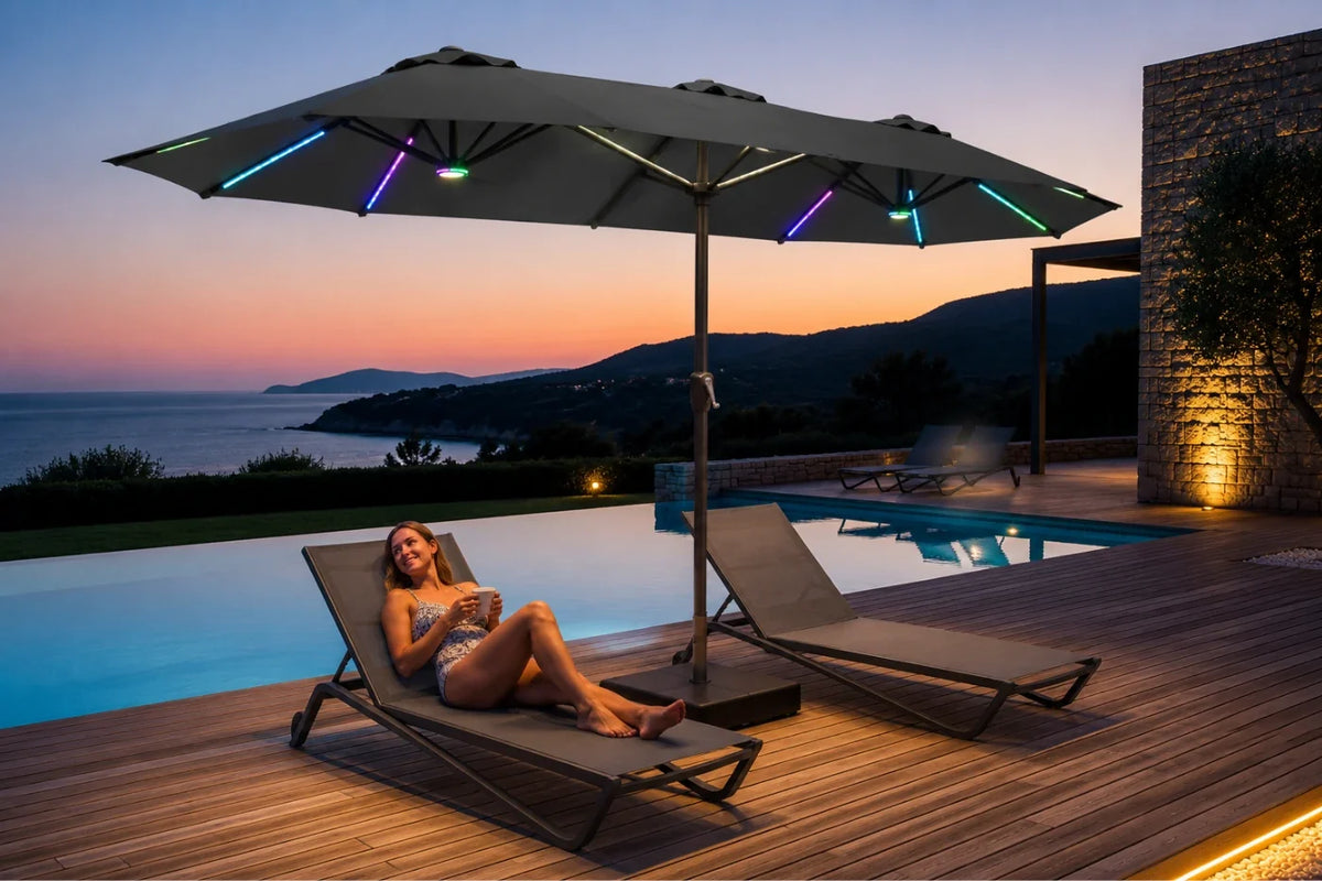A large dark gray patio umbrella with integrated multicolor RGB LED strip lights is open on a wooden deck beside an infinity pool at dusk, with a woman reclining on a lounge chair under the umbrella.
