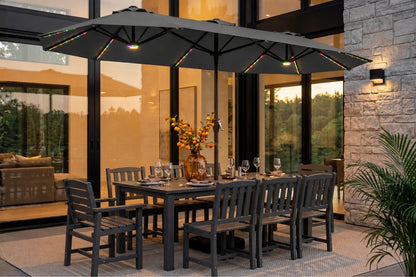 A dark gray outdoor dining set consisting of a rectangular table and 8 matching slatted chairs, styled under a matching large dark gray patio umbrella with integrated string lights on a stone patio.