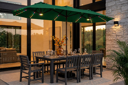 A dark green outdoor patio umbrella with integrated warm white string lights, shading a dark gray dining table and 8 matching chairs on a patio in front of a modern house.