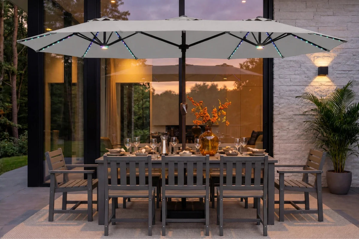 A large gray patio umbrella with integrated RGB LED string lights is shown over a dining table and chairs on a patio in front of a modern house at dusk.