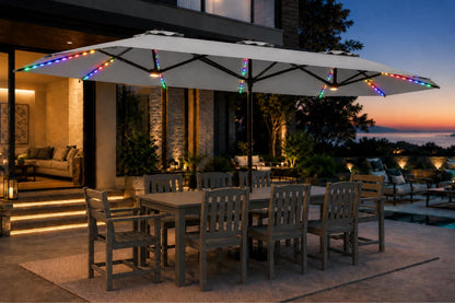A large gray patio umbrella with colorful RGB LED string lights along the canopy edges, shading an outdoor dining table and chairs on a patio at dusk.