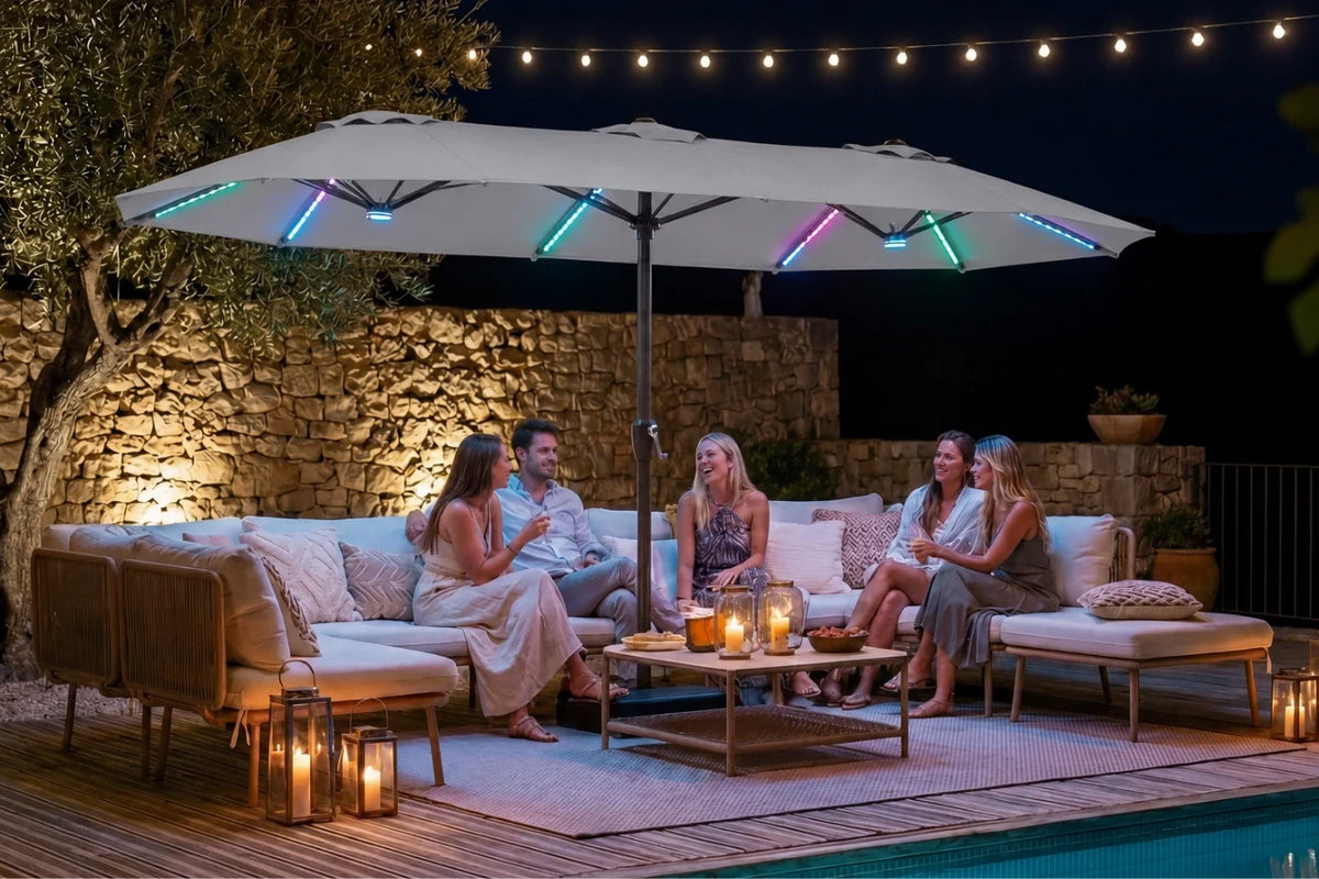 A large gray patio umbrella with integrated RGB LED lighting is set up on a wooden deck at night, shading a group of five people sitting on a beige outdoor sofa. The scene is illuminated by warm string lights, candlelight, and the colorful changing lights on the umbrella canopy.