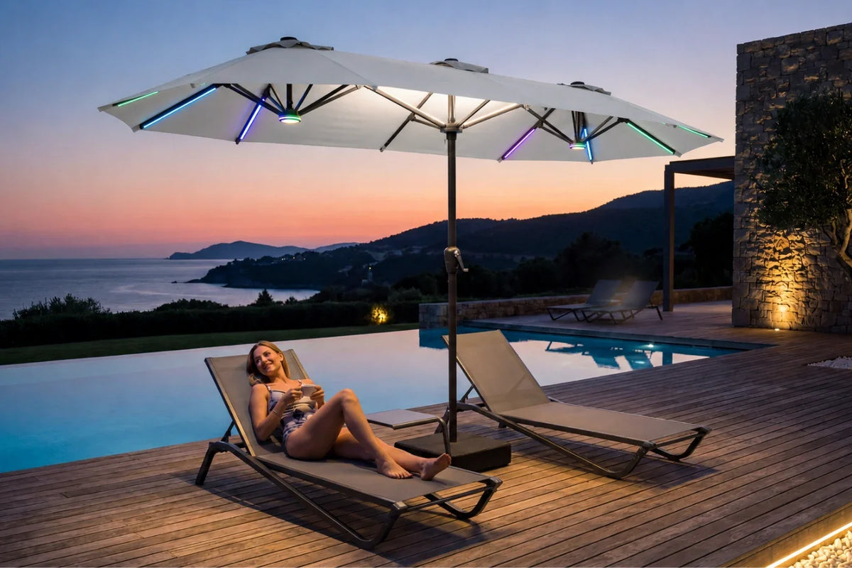 A woman relaxes on a beige lounge chair beside a gray outdoor patio umbrella with integrated RGB LED lighting, set next to an infinity pool on a wooden deck at dusk.