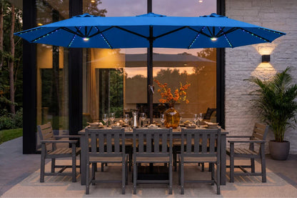 A large indigo blue patio umbrella with integrated string lights is shown over a gray outdoor dining table and chairs on a patio at dusk, with a modern house interior and exterior visible in the background.