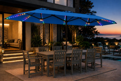 A large indigo blue outdoor patio umbrella with multicolor RGB LED string lights draped along the canopy and frame, set on a patio at dusk next to a dining table and chairs.