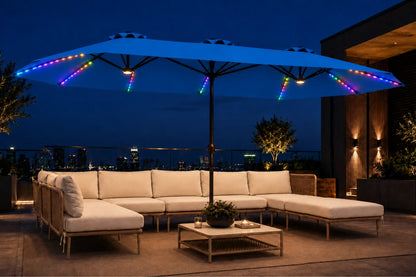 Outdoor patio furniture set under a large indigo blue patio umbrella with colorful RGB LED string lights, shown at night on a rooftop terrace with a city skyline in the background.