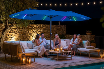 A large indigo blue patio umbrella with colorful RGB LED light strips is set over an outdoor lounge area at night, with four people sitting underneath on a beige sectional sofa. The scene is illuminated by warm string lights, candles, and the blue umbrella, with a stone wall and pool visible in the background.