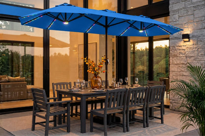 A indigo blue patio umbrella with integrated RGB LED lighting is shown outdoors over a dark gray dining table and chairs on a patio in front of a modern glass house.
