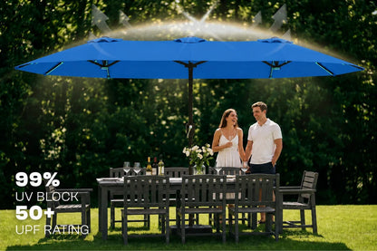 A large indigo blue patio umbrella with a built-in misting sprayer spraying water overhead, shading a dark outdoor dining table and chairs. A couple stands under the umbrella in a sunny backyard with trees in the background.