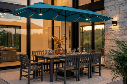 A lake blue outdoor patio umbrella with integrated RGB LED string lights is shown over a dark gray rectangular dining table and matching chairs on a patio in front of a modern house.