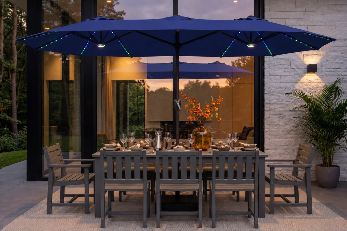 A large navy blue patio umbrella with integrated RGB LED string lights is shown open over a gray outdoor dining table and chairs on a patio. The setting is a modern house exterior at dusk with glass windows, a stone wall, and a potted plant.