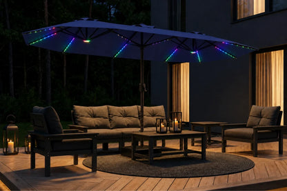 Outdoor patio furniture set under a large navy blue patio umbrella with multicolor RGB LED string lights at night, featuring a sectional sofa, two armchairs, and a coffee table on a wooden deck.