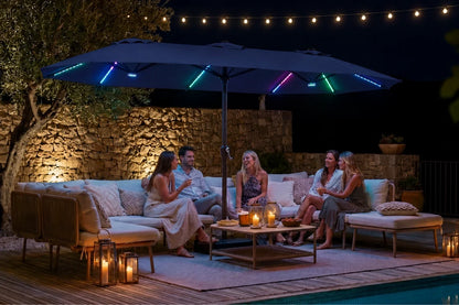 Outdoor patio scene at night with a large navy blue patio umbrella illuminated by colorful RGB LED light strips, surrounded by a lounge sofa, people sitting and chatting, candles, and string lights.