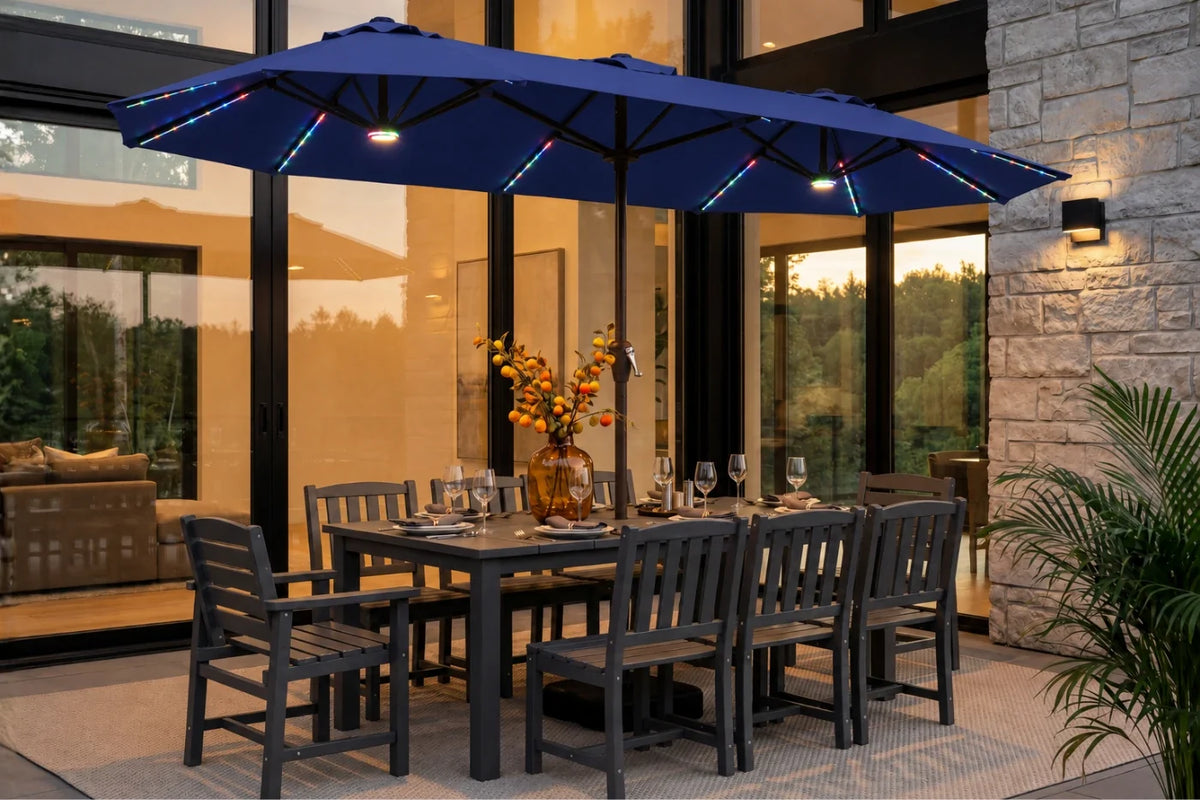 A navy blue outdoor patio umbrella with integrated RGB LED string lights is shown over a dark gray dining table and chairs on a patio. The setting is a modern home patio at dusk with stone walls, glass windows, and a potted plant in the foreground.
