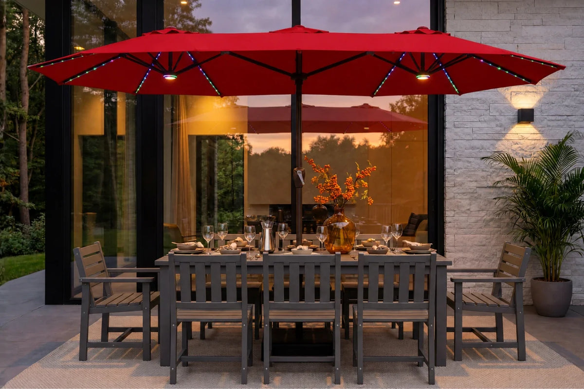 A red patio umbrella with integrated string lights over a gray outdoor dining table and chairs on a patio in front of a modern glass house at dusk.