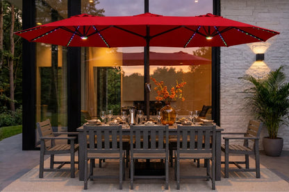 A red patio umbrella with integrated string lights over a gray outdoor dining table and chairs on a patio in front of a modern glass house at dusk.