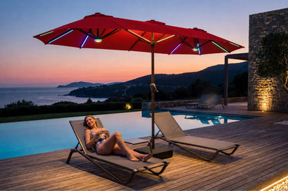 A woman relaxes on a beige lounge chair beside a pool at dusk, under a large red patio umbrella with colorful RGB LED light strips. A second lounge chair sits nearby on a wooden deck, with a stone wall and distant hills across the water in the background.