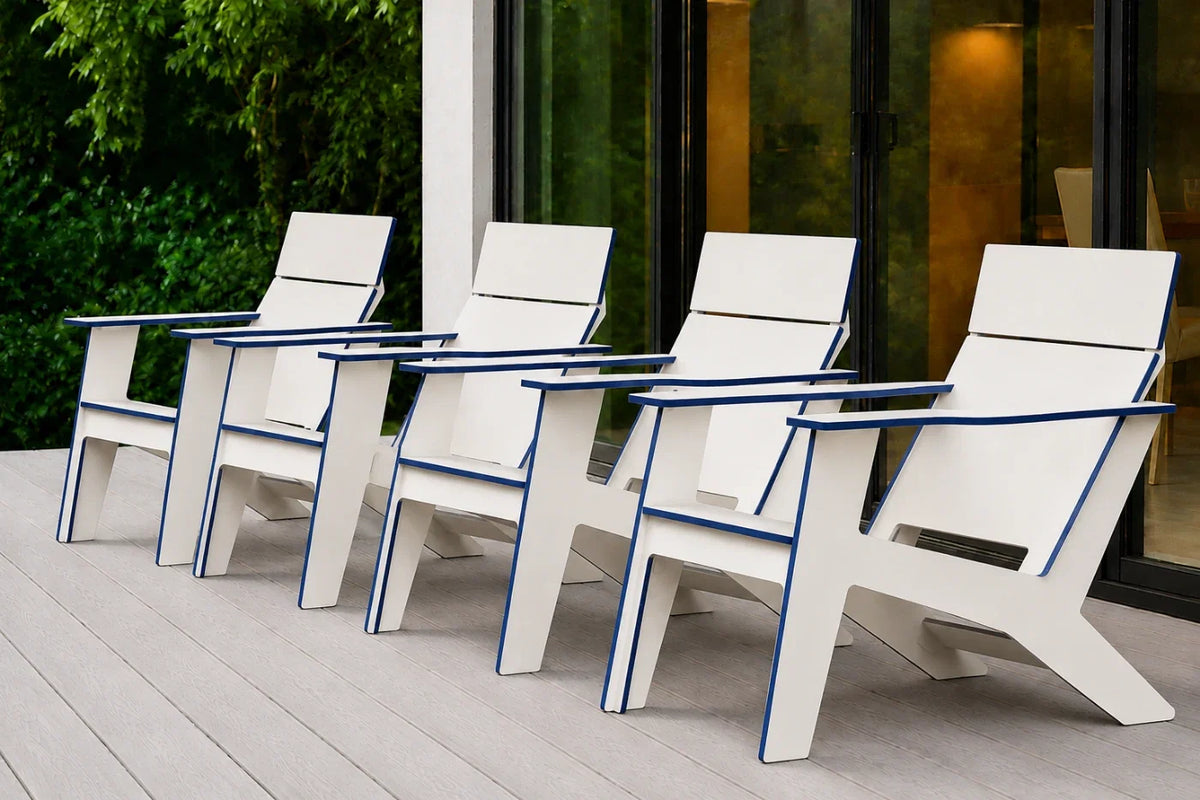 Four white outdoor Adirondack chairs with blue trim arranged in a row on a light gray deck in front of a modern glass building with greenery in the background.