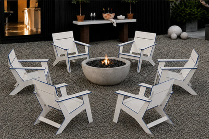 Six white outdoor Adirondack-style chairs arranged in a circle around a round concrete fire pit on a gravel patio, with a black wall, table, plants, and decorative spheres in the background.