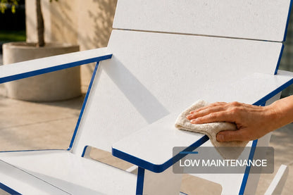 A close-up of a white modern poly Adirondack chair with blue trim being wiped with a cloth by a person's hand, with a 'LOW MAINTENANCE' text overlay.
