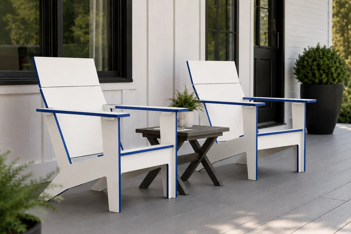 Two modern white outdoor Adirondack chairs with blue trim are shown on a gray porch next to a small dark side table. The background features a white house exterior with black-framed windows and a potted green shrub.