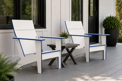 Two modern white outdoor Adirondack chairs with blue trim are shown on a gray porch next to a small dark side table. The background features a white house exterior with black-framed windows and a potted green shrub.
