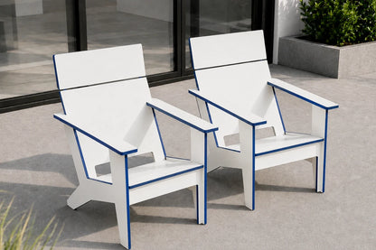 Two modern outdoor Adirondack-style chairs in white with blue trim, shown on a concrete patio in front of a glass door.