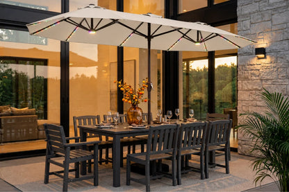 Outdoor dining set with a rectangular dark gray table, 8 matching chairs, and a large creamy white patio umbrella with integrated string lights, staged on a patio in front of a modern house.