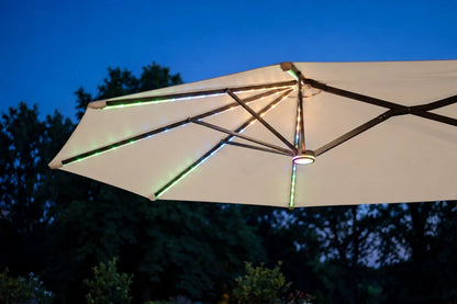 A large patio umbrella with a creamy white fabric canopy and dark metal frame, shown outdoors at dusk with integrated RGB LED lighting glowing along the ribs and canopy edges against a dark blue sky and trees.