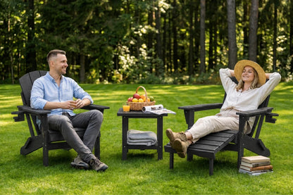 A man and a woman relax in black outdoor Adirondack chairs on a grassy lawn with a small table between them, set against a dense green forest background.